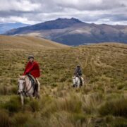 Image of two students in ponchos riding horses over grassy hill away from mountains in the distance
