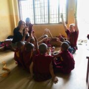 Students sits with young kids in circle on the floor getting them to raise their arms, an adult takes a photo on her camera