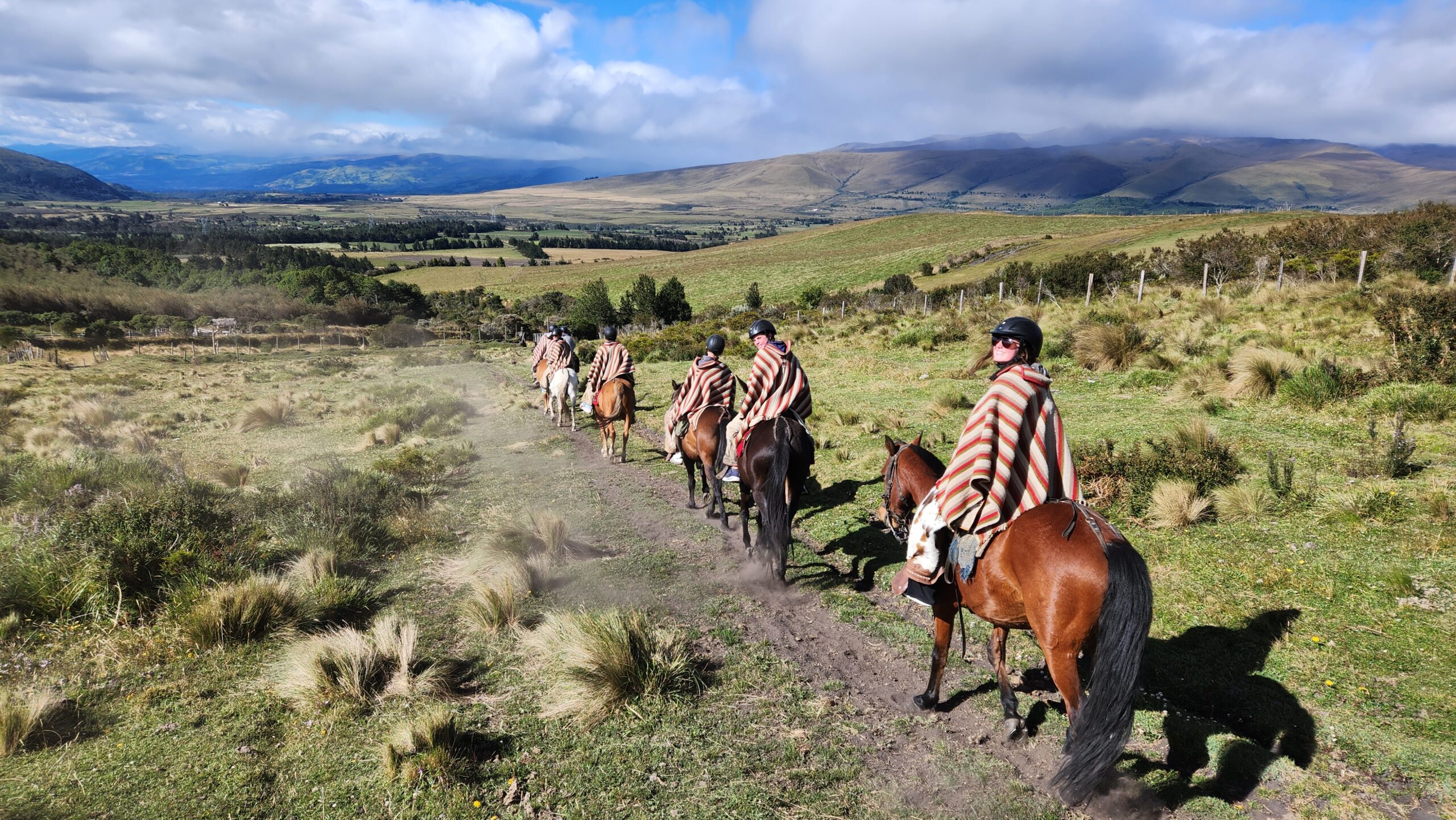 Students in striped ponchos ride in a line on horses towards mountain range