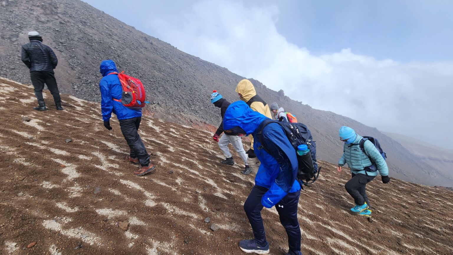 Students in colorful wind gear hike up rocky slope of Cotopaxi