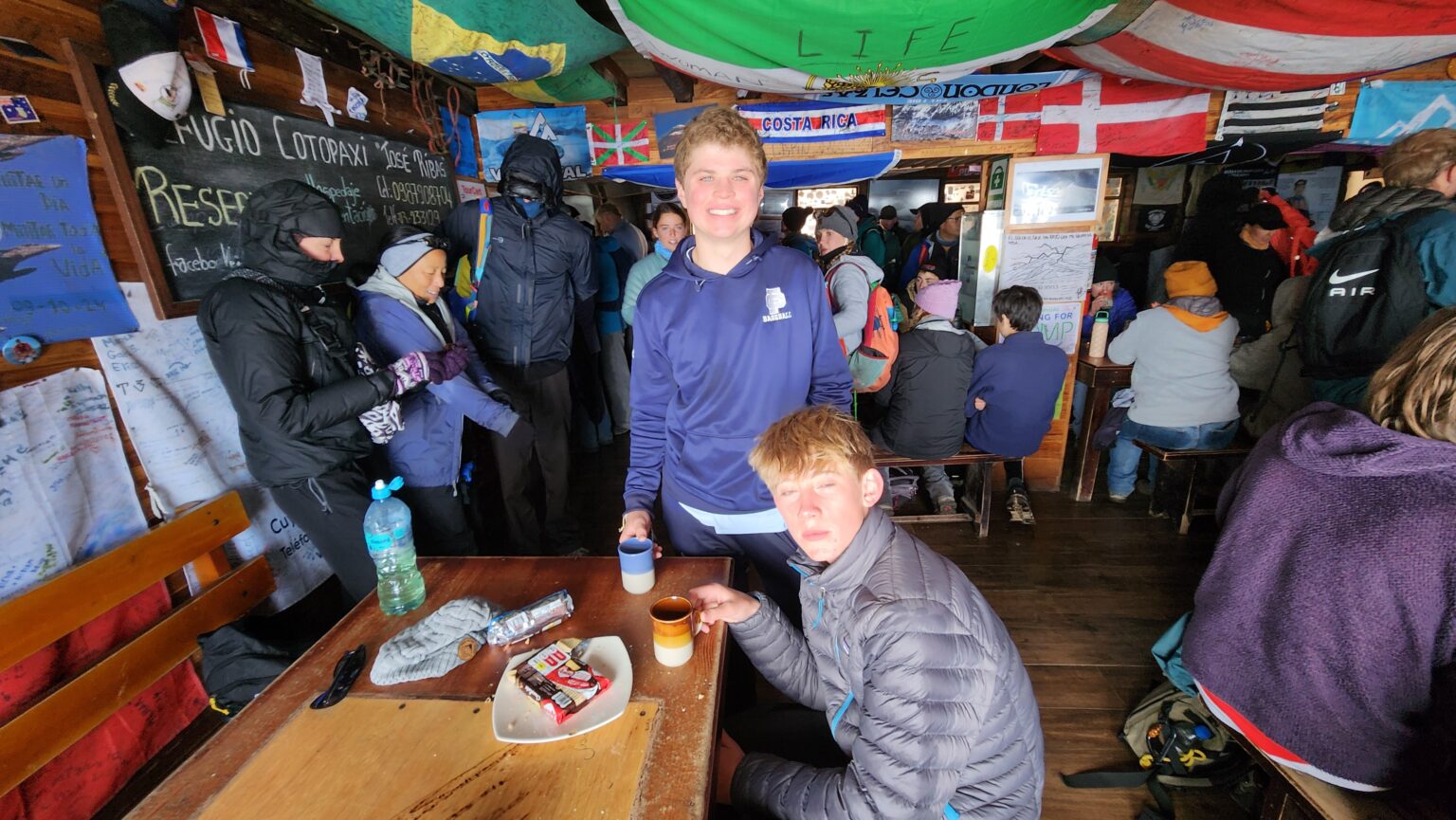 Two students sit inside small building with many colorful flags drinking warm beverages 
