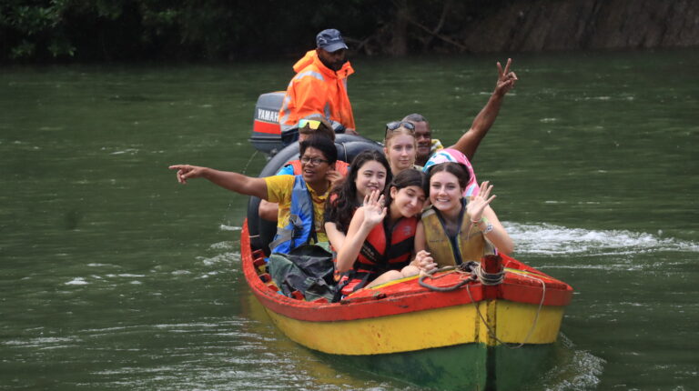 Students sit in boat, many pointing, voyaging down the Navua River in the Fijian jungle.