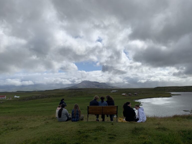 Students sit on a bench in a grassy field looking out at mountains and lake