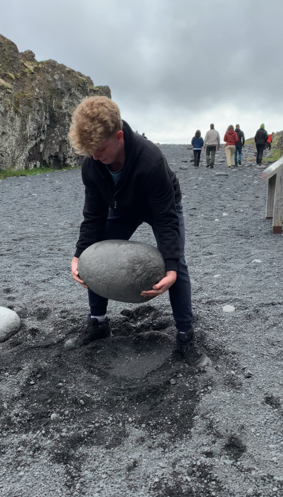 Student tries to life large stone on black sand beach