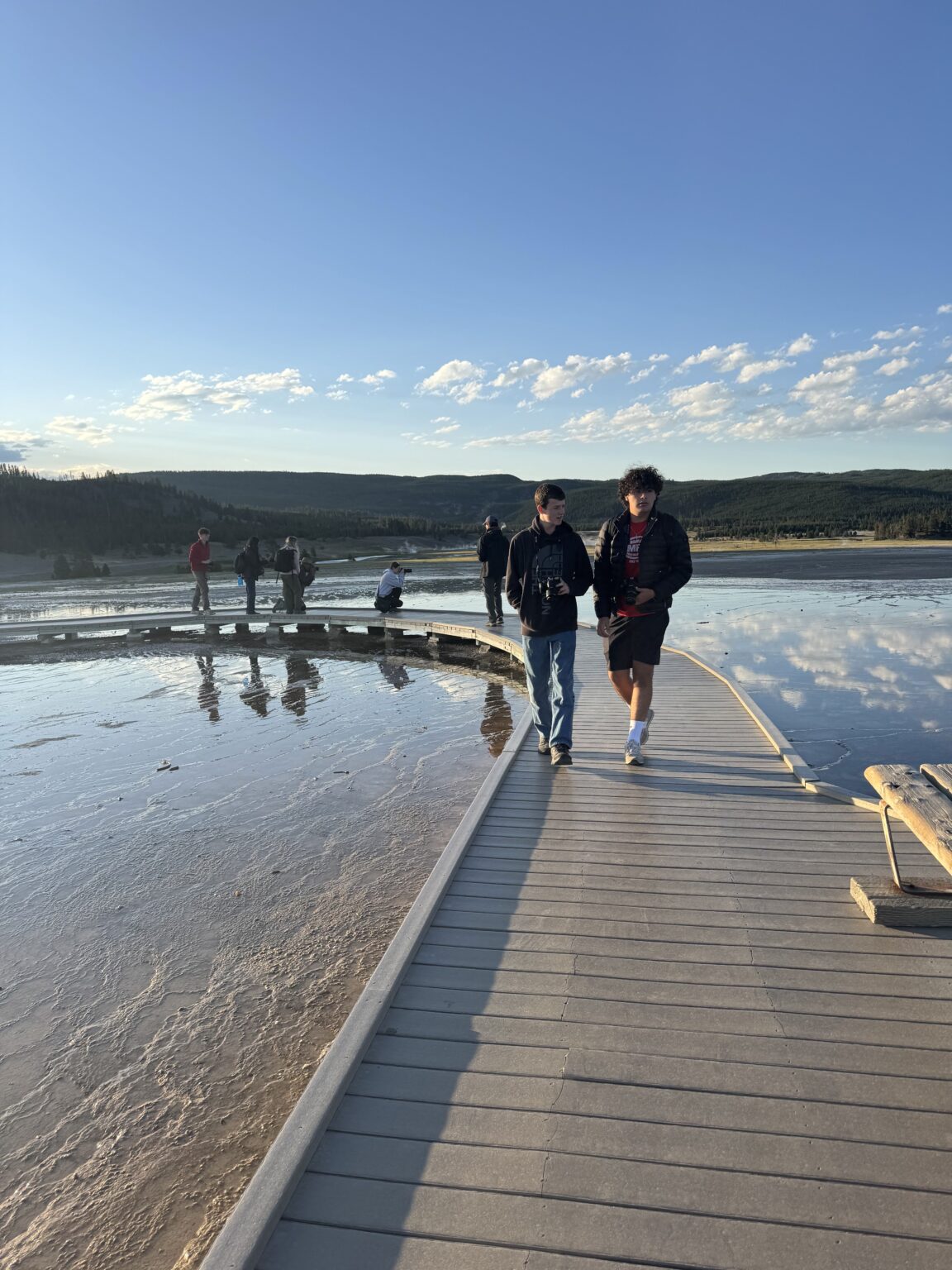 Students walk on bridge with water reflecting blue sky, clouds, and mountain range behind