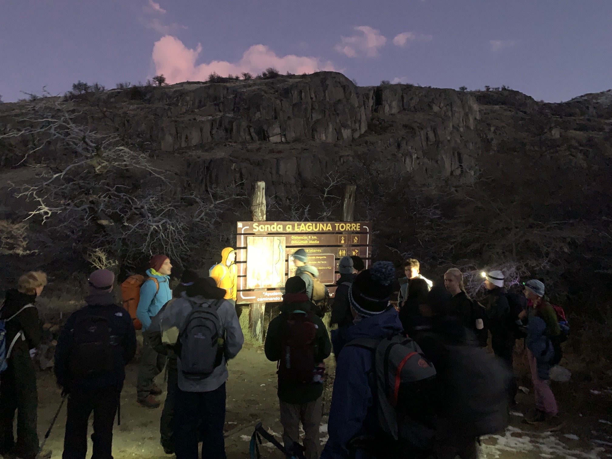 Students stand outside at dawn looking at mountain trail map on signage board