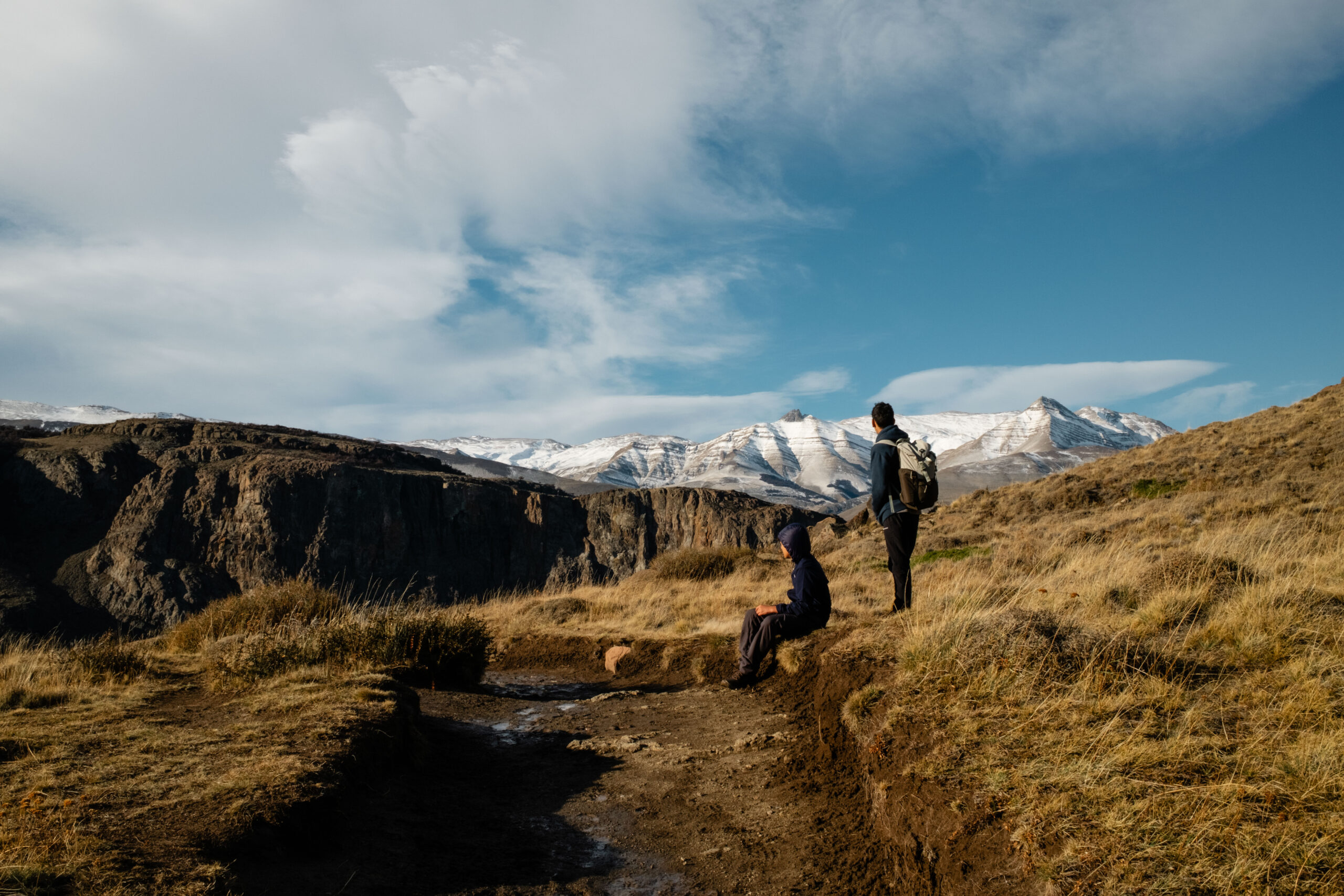 Two students sit and stand by a dirt road looking out at snowy mountains in the distance