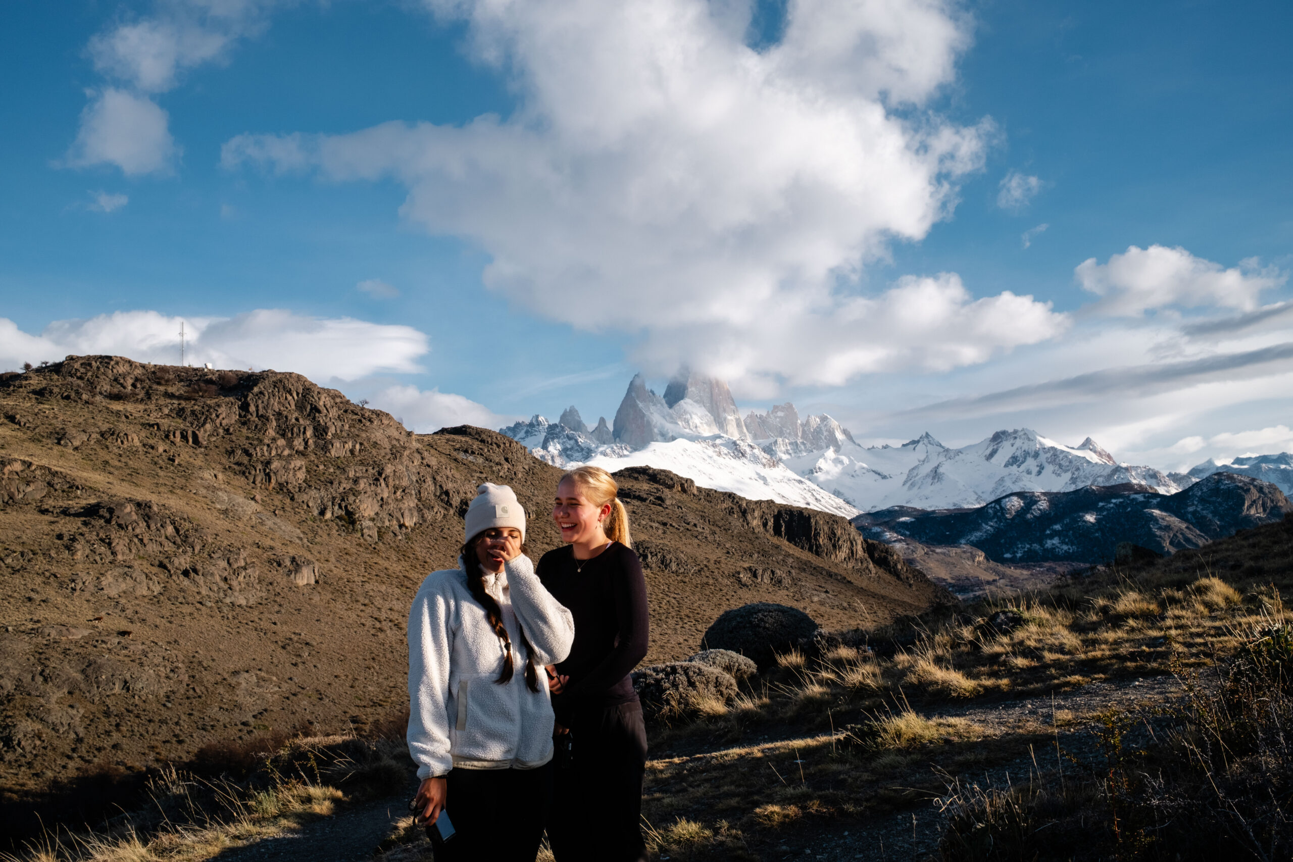 Two students laugh posing for a picture with snowy mountain peaks visible in the distance