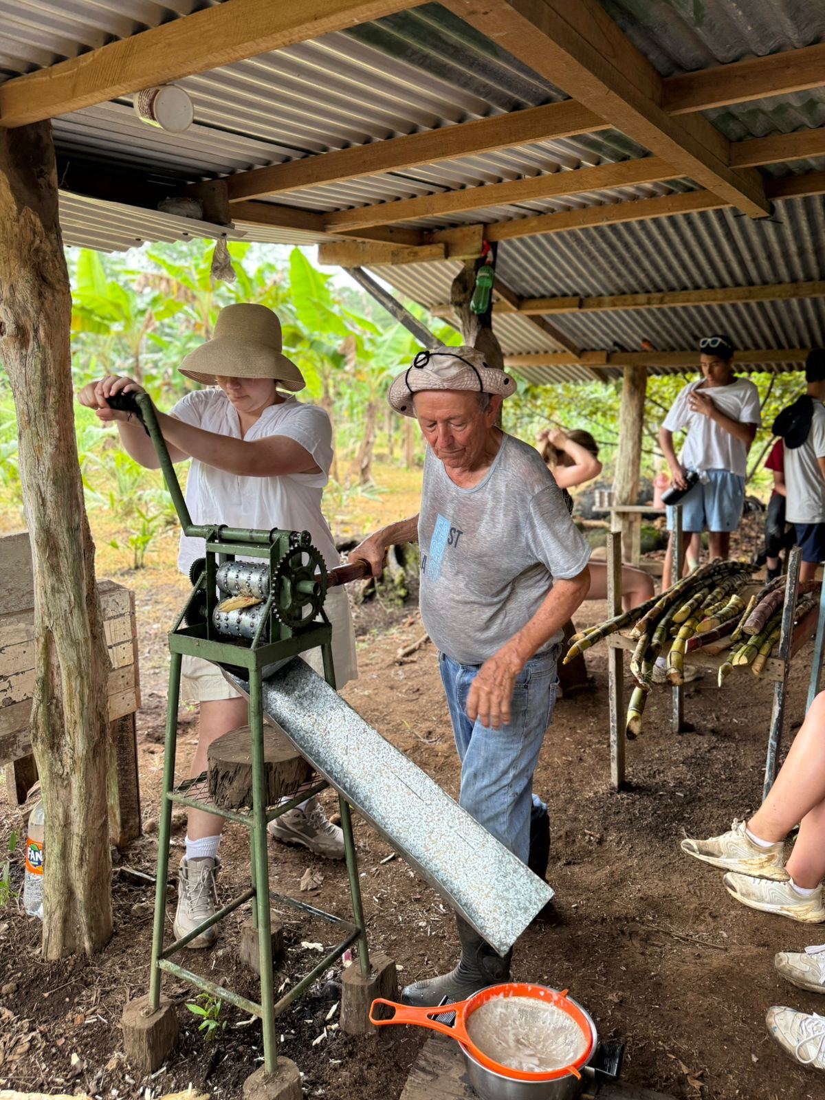 Local instructor lets student crank metal machine to collect sugarcane juice that slides down metal pan