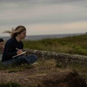 Student sits on grass, wind blowing back hair, overlooking hills with notebook in hand
