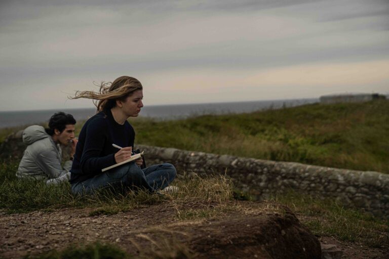 Student sits on grass, wind blowing back hair, overlooking hills with notebook in hand