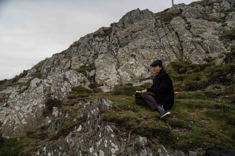 Student sits on grassy patch with a notebook, rocks an cliffs behind them