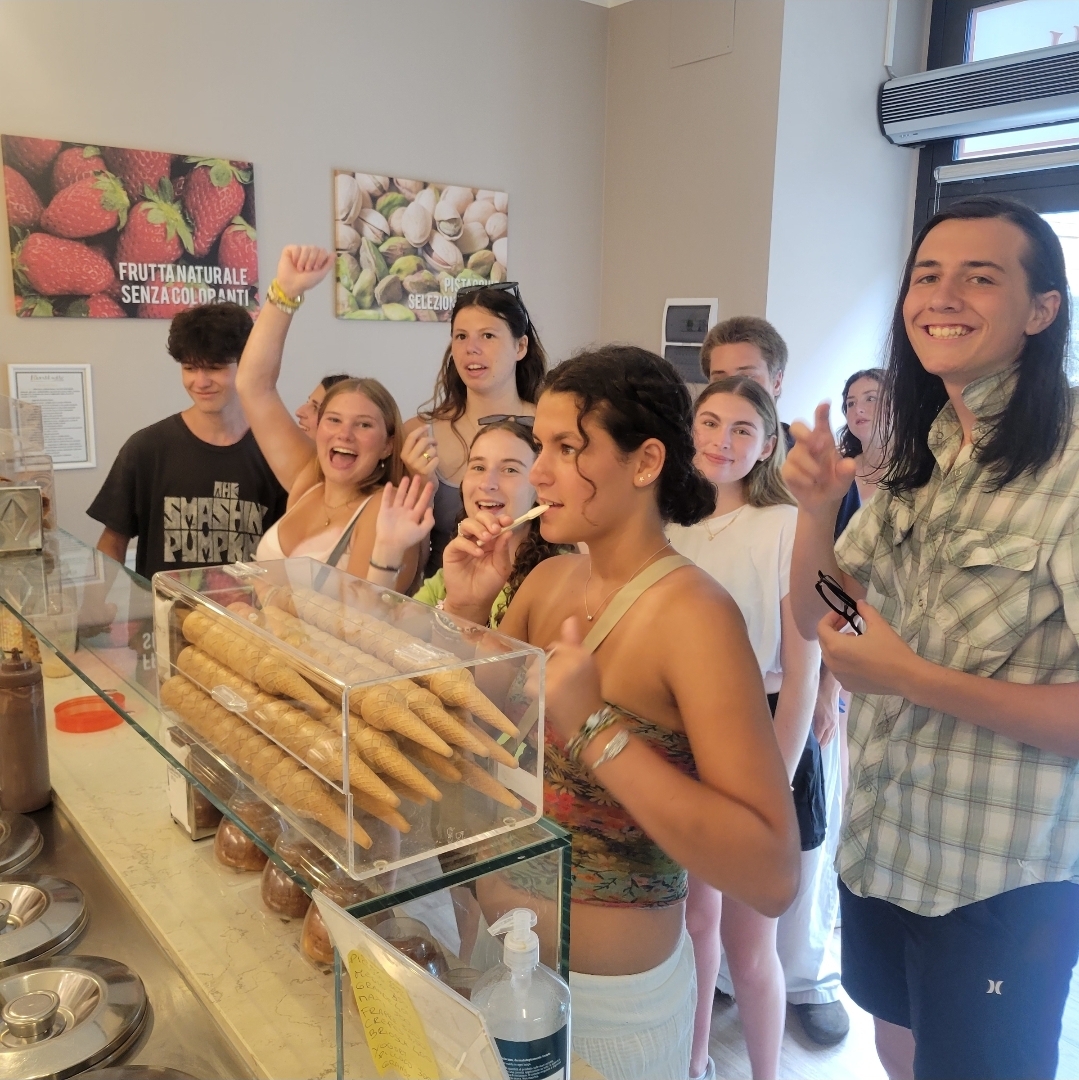 Student stand behind a gelato counter, one student hold small spoon tasting a flavor