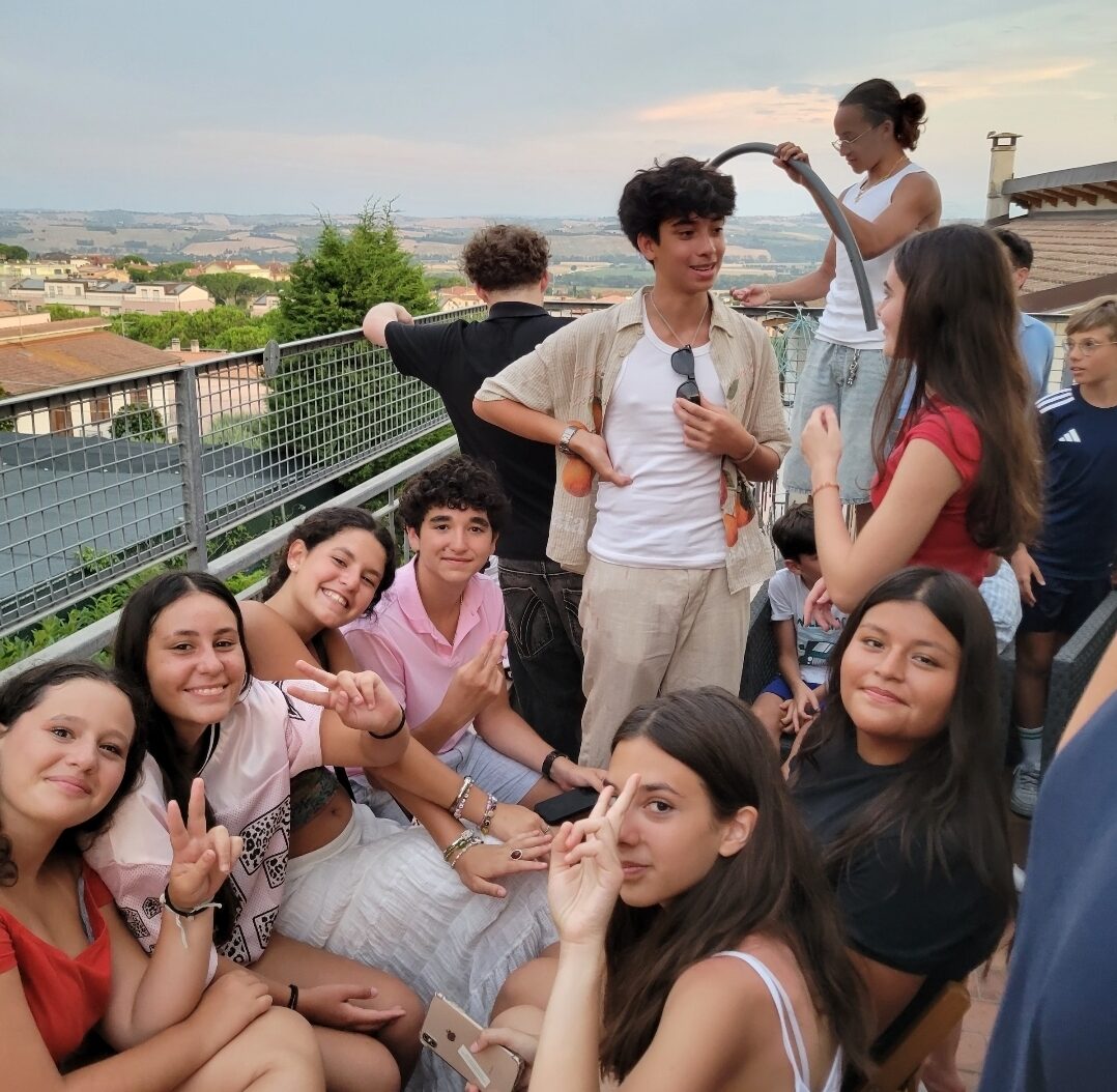 Student sit and stand near tables, some hold up peace signs, posing for picture, Italian city of Osimo can been seen in background