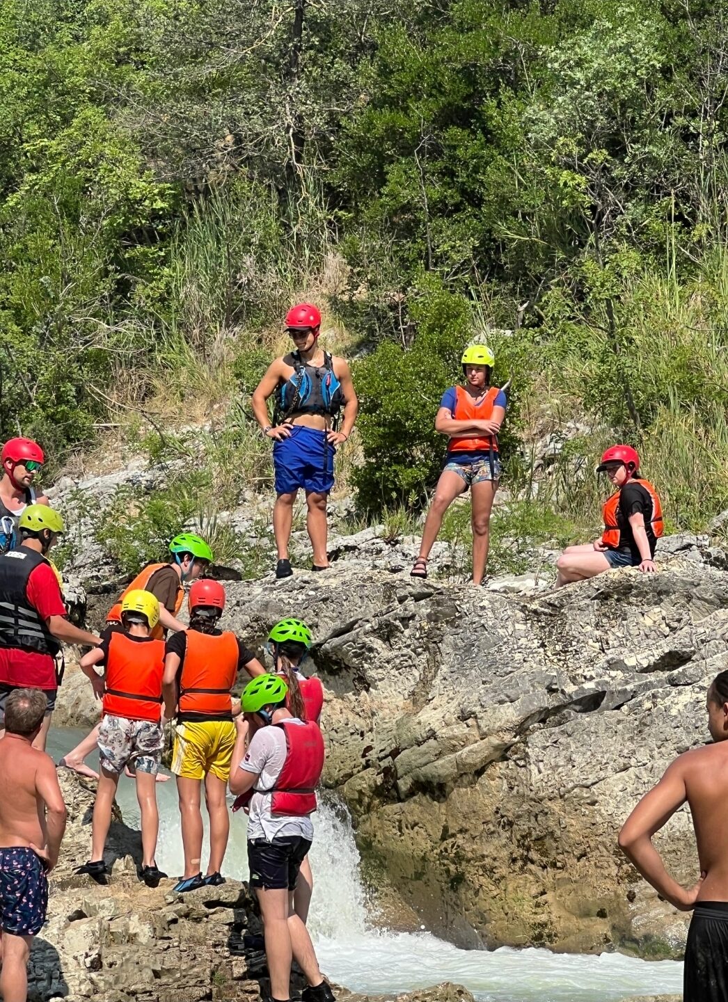 Students climb rocks outside near riverside wearing life jackets and helmets