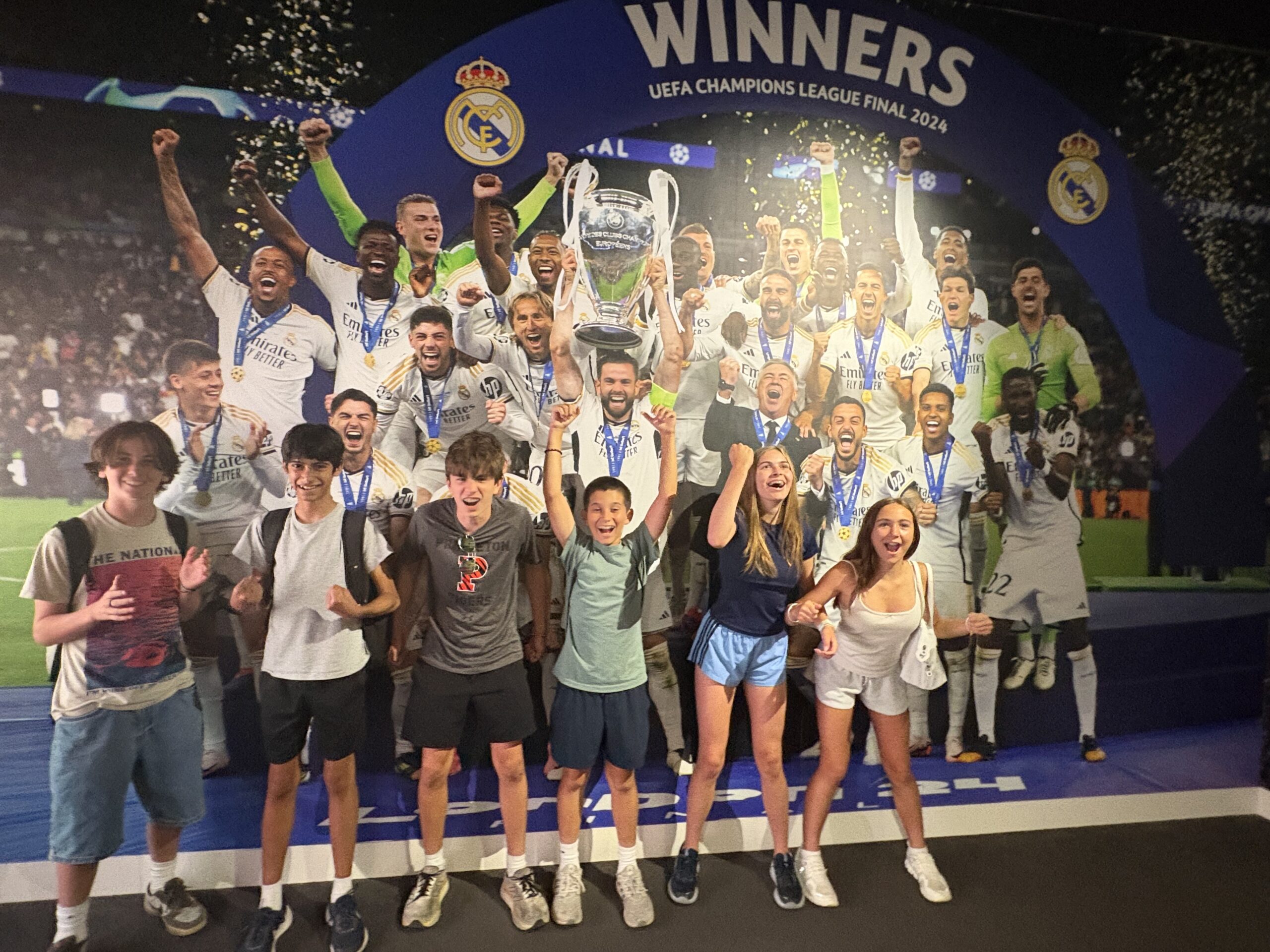 Students pose for a group photo hands up next to a sign of a past soccer winners