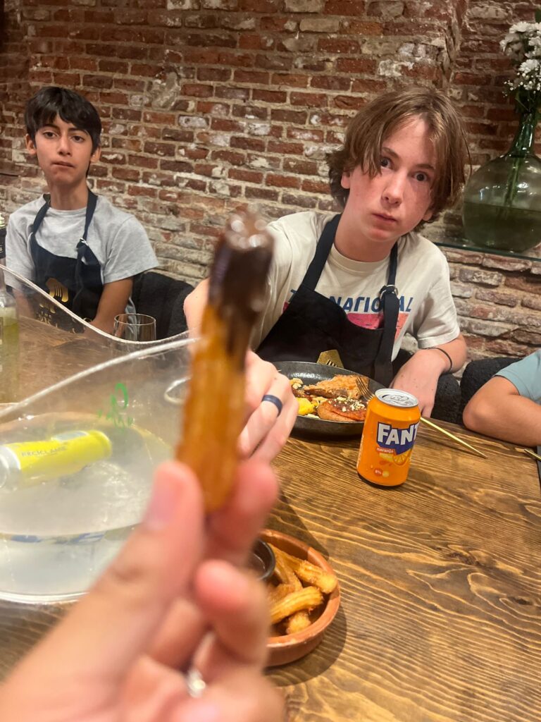 Someone holds up a churro covered in chocolate while two students, eyes wide, gaze at it
