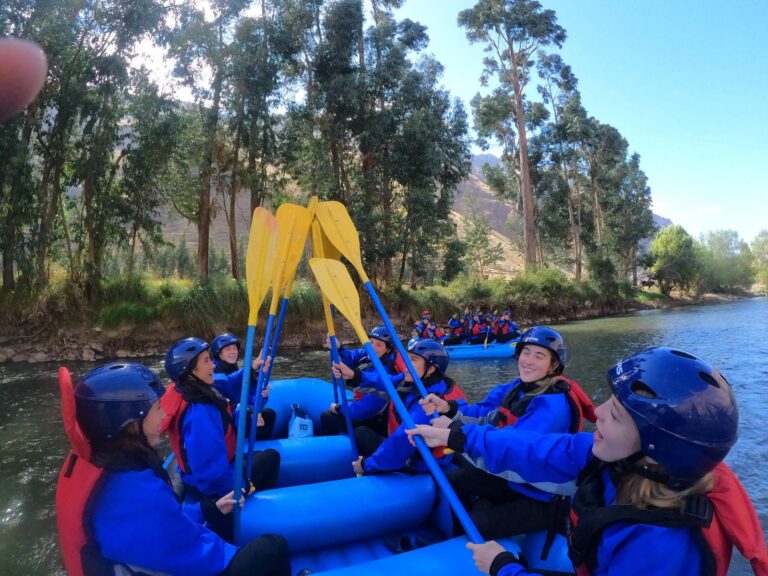 Students on a raft wearing blue safety gear raise their oars together in a cheer